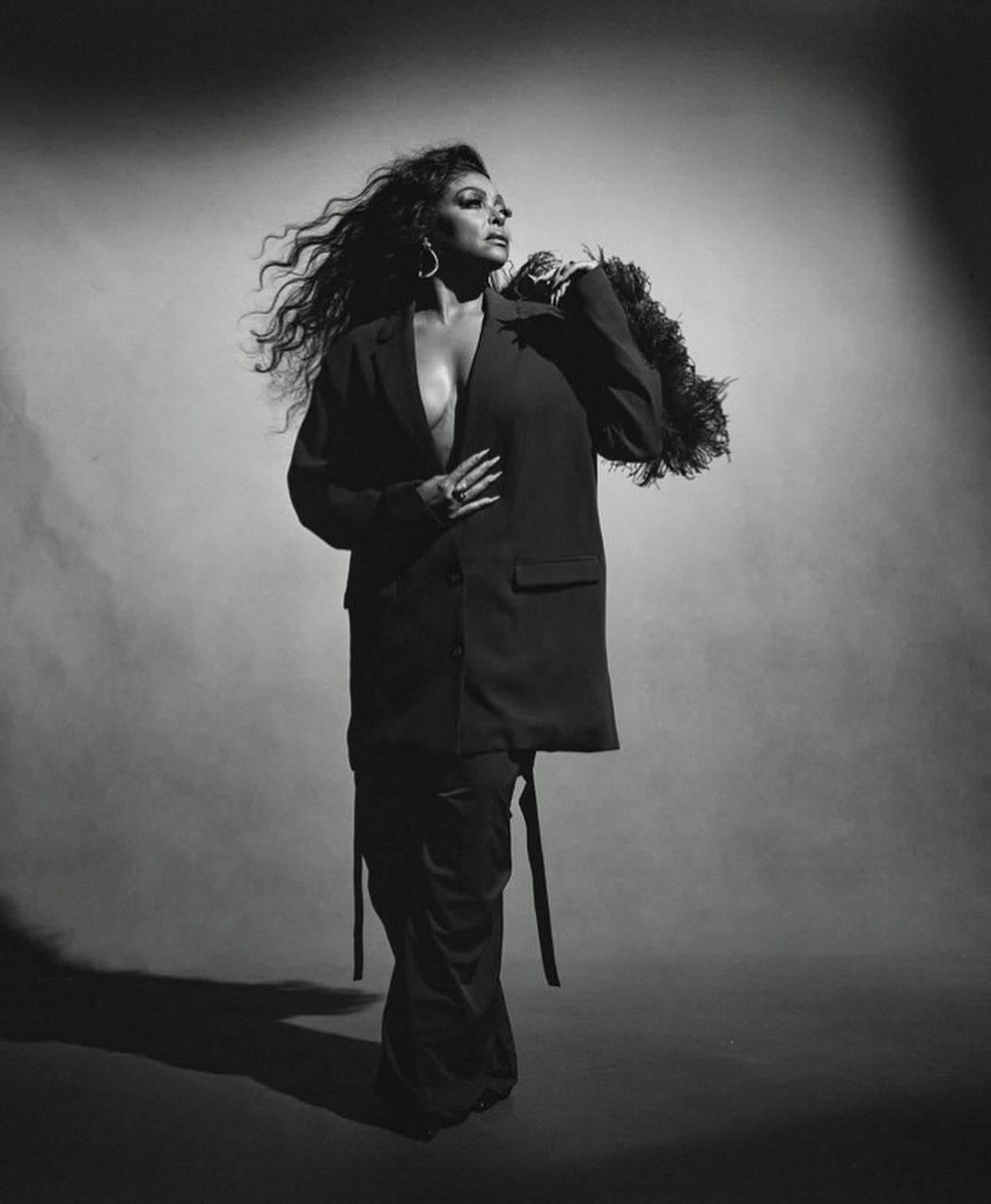 Black and white portrait of a woman with long wavy hair wearing an oversized blazer and holding a feather piece by Kanya Iwana.