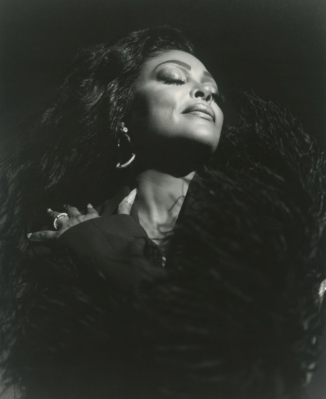 Black and white portrait of an African American woman with long curly hair wearing jewelry and a feathered shawl against a dark background.