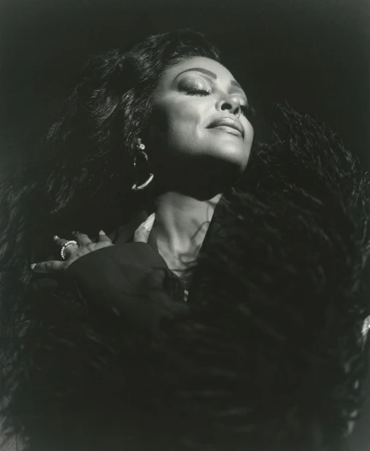 Black and white portrait of an African American woman with long curly hair wearing jewelry and a feathered shawl against a dark background.