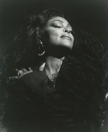 Black and white portrait of an African American woman with long curly hair wearing jewelry and a feathered shawl against a dark background.