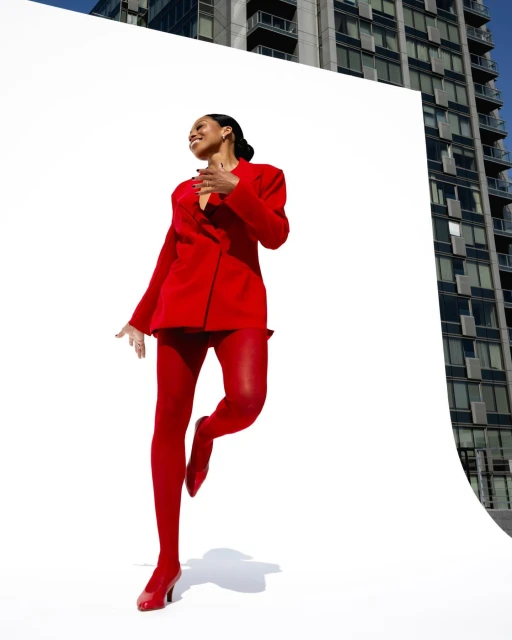 A smiling woman in a red suit and tights poses in front of a white backdrop, with a tall building visible in the background on a sunny day.
