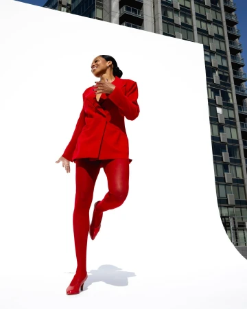 A smiling woman in a red suit and tights poses in front of a white backdrop, with a tall building visible in the background on a sunny day.