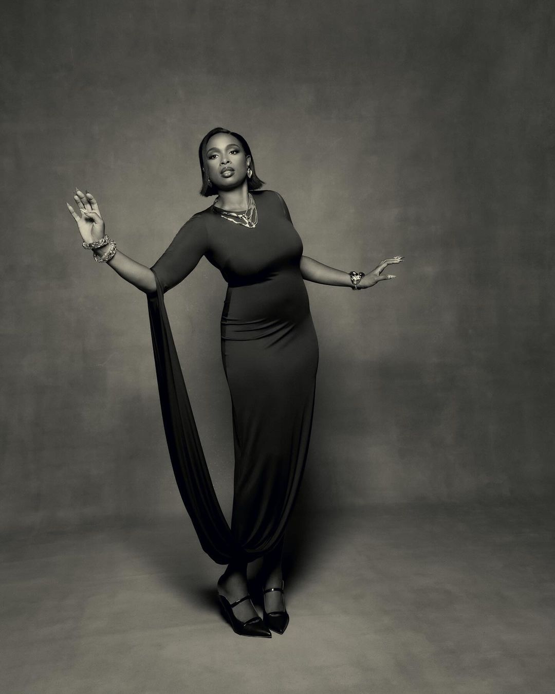 Monochrome portrait of a woman in a long fitted dress with statement jewelry against a textured backdrop
