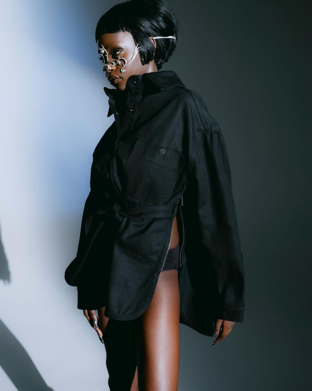 Woman with dark hair and a jeweled mask poses in a black jacket with a belt. The model is in a studio environment with soft lighting.