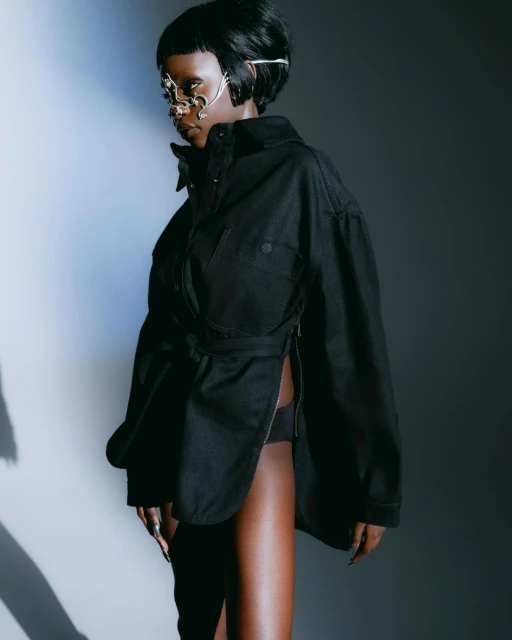 Woman with dark hair and a jeweled mask poses in a black jacket with a belt. The model is in a studio environment with soft lighting.