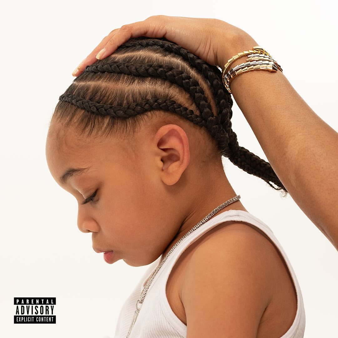 Profile view of child with cornrow braids and silver chain, adult hand atop their head, wearing bracelets, against a white backdrop.