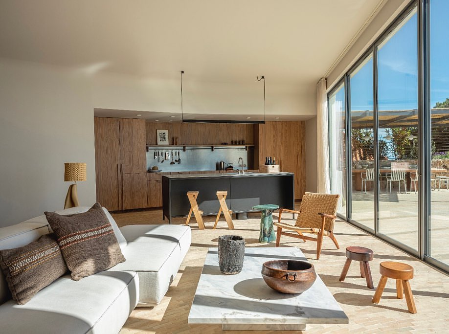 Living room interior with white couch, marble coffee table, stools, and open kitchen in the background, bathed in natural light.