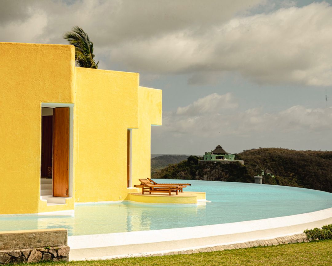 Yellow building overlooks an infinity pool and a distant hillside. Outdoor lounge chairs sit near the water's edge, with a green building in the background.