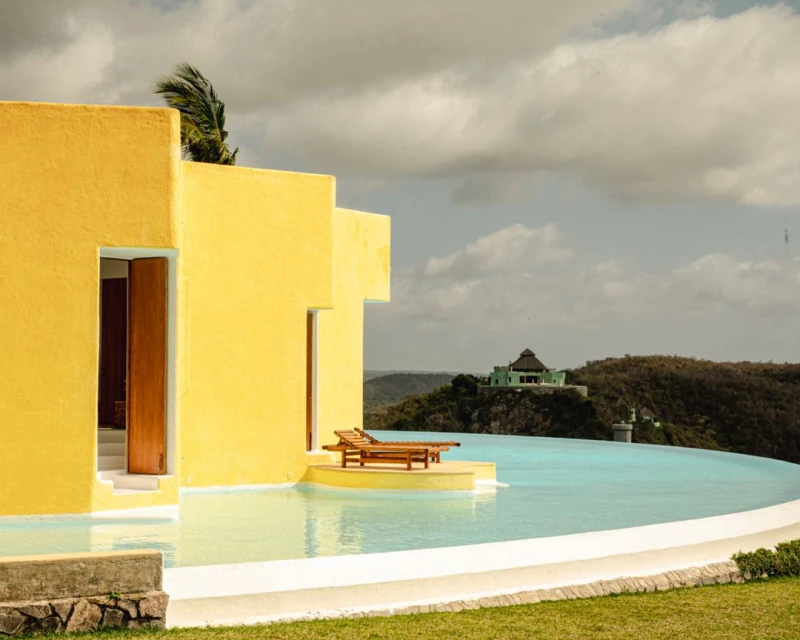 Yellow building overlooks an infinity pool and a distant hillside. Outdoor lounge chairs sit near the water's edge, with a green building in the background.