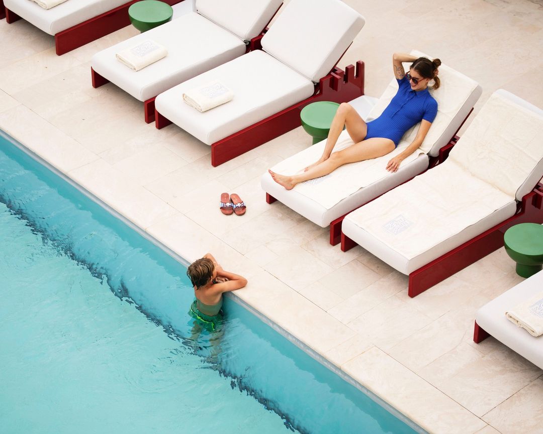 Overhead shot of woman in swimsuit relaxing on lounge chair near a pool with a child in the water at a resort on a sunny day.
