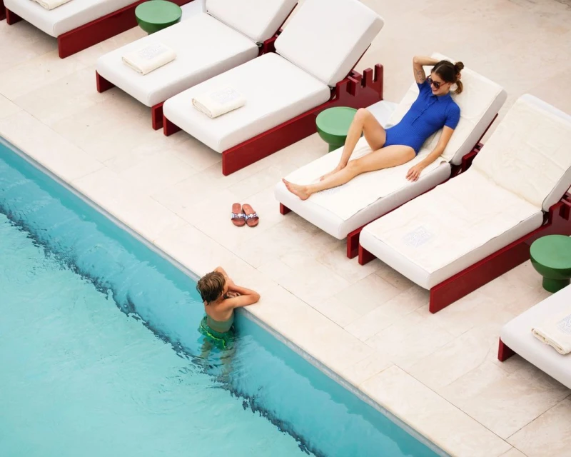 Overhead shot of woman in swimsuit relaxing on lounge chair near a pool with a child in the water at a resort on a sunny day.