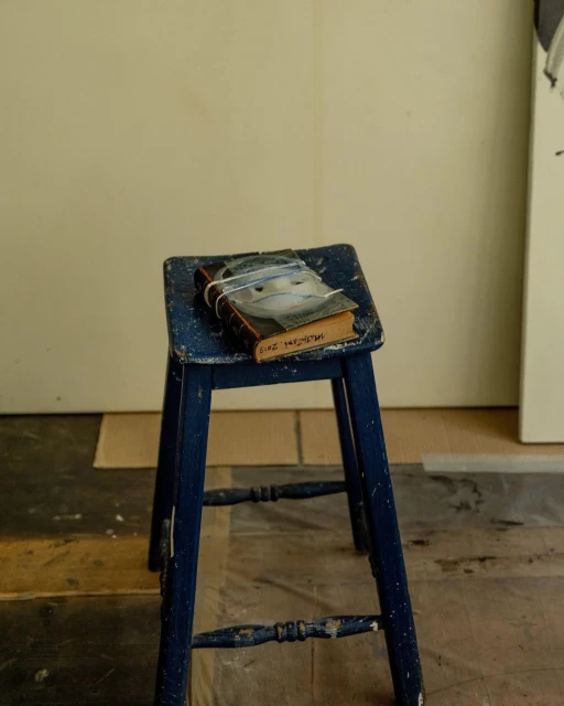 Blue painted wooden stool with book featuring face image on top in indoor setting with warm lighting, textured floors, and neutral wall.