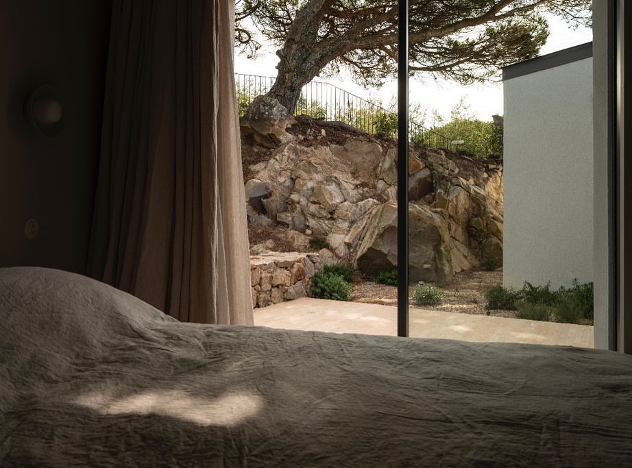 Bedroom view through large glass window to an outdoor stone patio and boulder landscape with a leafy tree and white building.