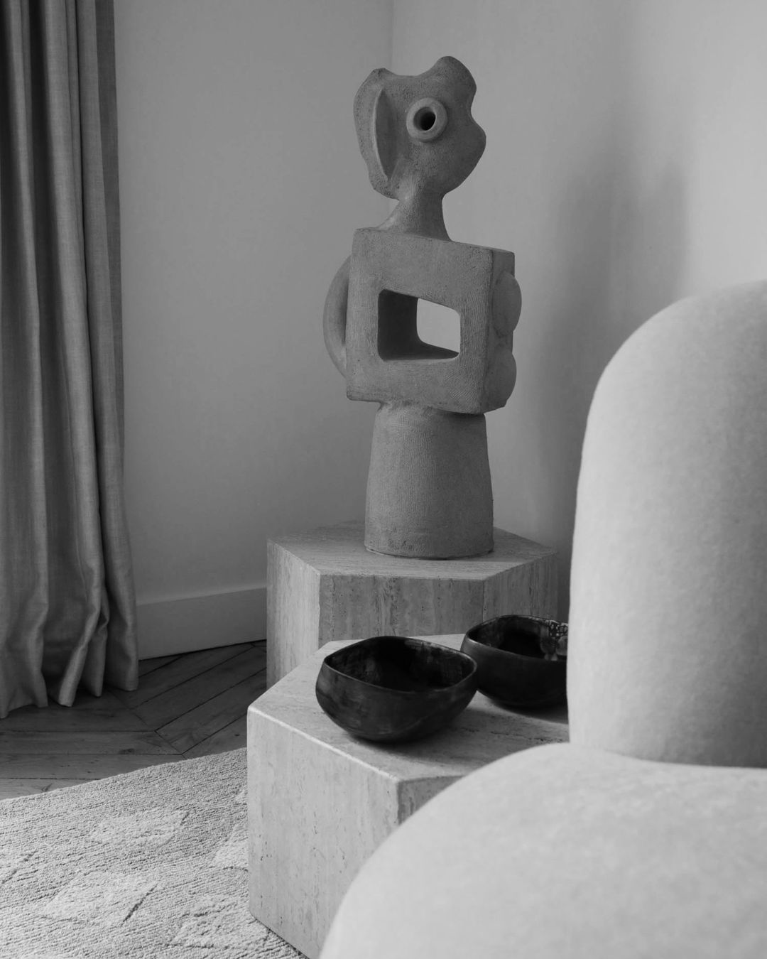 Contemporary interior features an abstract sculpture on a hexagonal pedestal table alongside a neutral-toned sofa and two bowls.