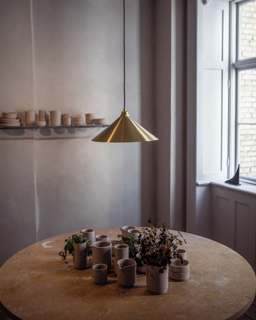 Still life arrangement featuring an overhead gold light fixture illuminating a round table with an assortment of ceramic vases and foliage.