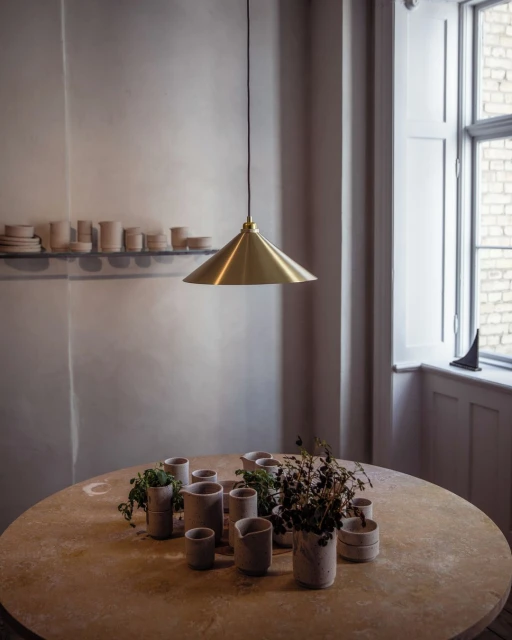 Still life arrangement featuring an overhead gold light fixture illuminating a round table with an assortment of ceramic vases and foliage.