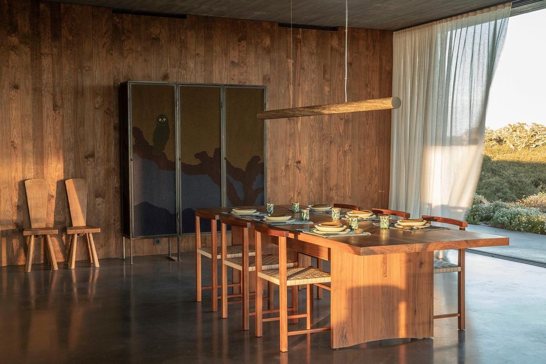 Dining area features a wooden table set for a meal, surrounded by wooden chairs, in a room with wooden walls and natural light.