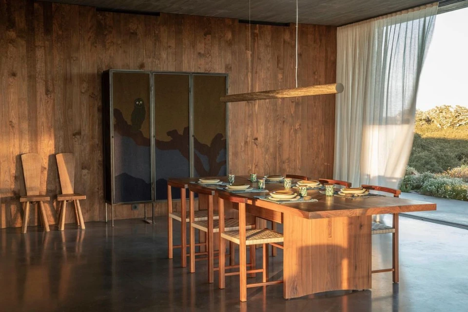 Dining area features a wooden table set for a meal, surrounded by wooden chairs, in a room with wooden walls and natural light.