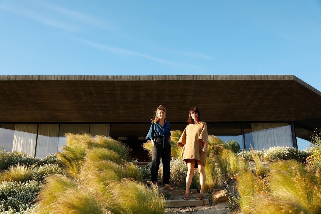 Two women stand on steps in front of a modern house with lush green plants under a clear blue sky. They are smiling and posing.