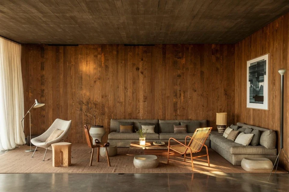 Interior view of a modern living room featuring a grey sofa, woven chair, wooden wall, and stone flooring, bathed in natural light.