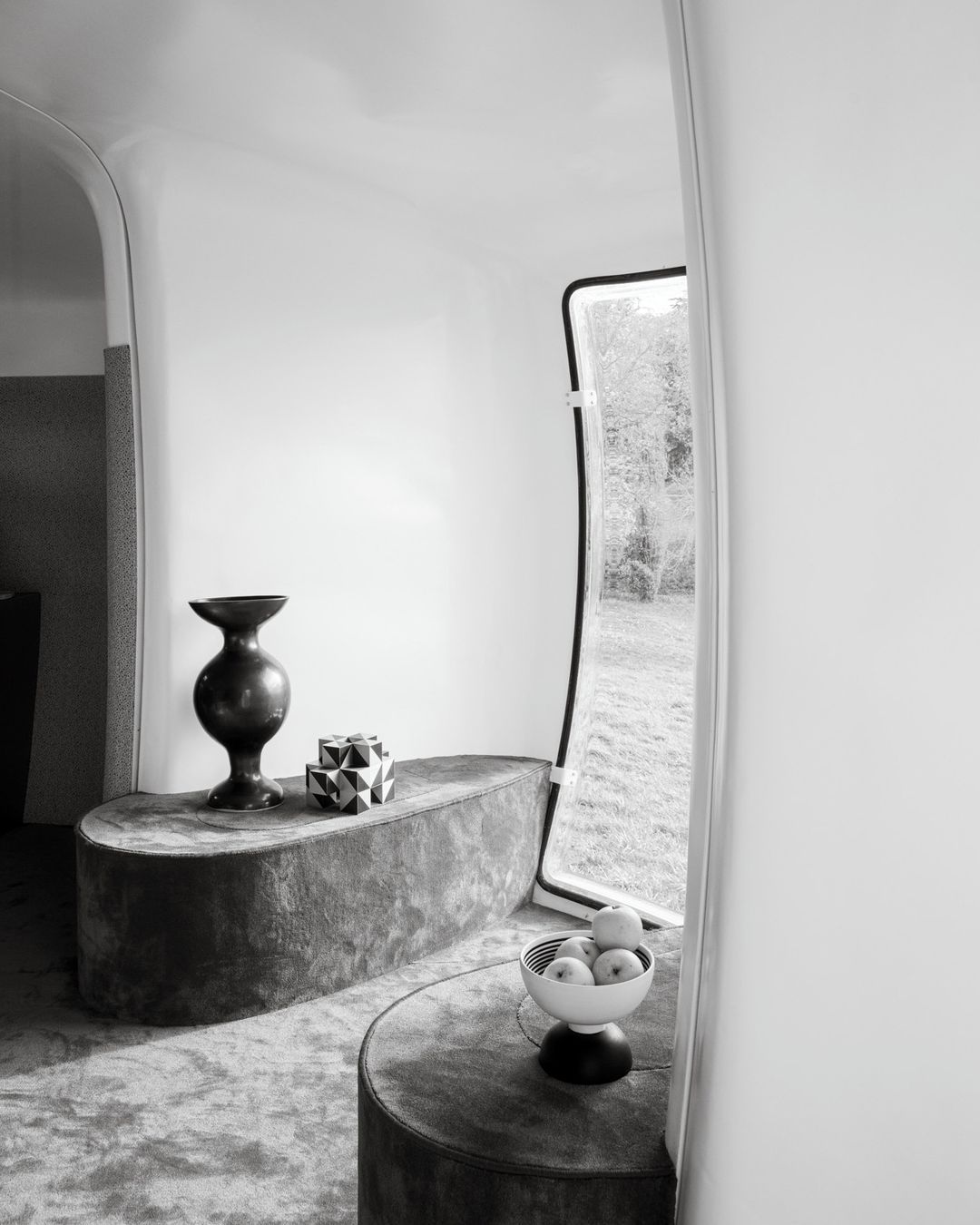 Modern interior space featuring a table with decorative vase, geometric figure, bowl of apples near a window, in monochrome.