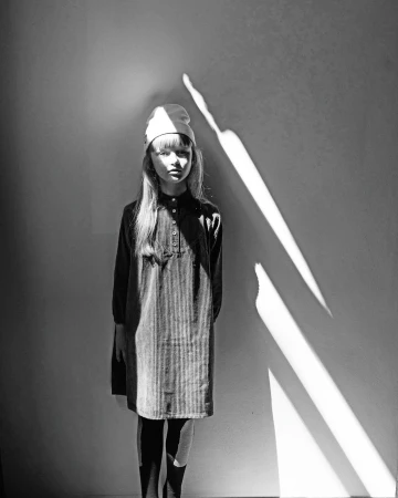 Monochrome portrait of a young girl wearing a beanie and dark button-down dress standing against a wall with strong diagonal light.