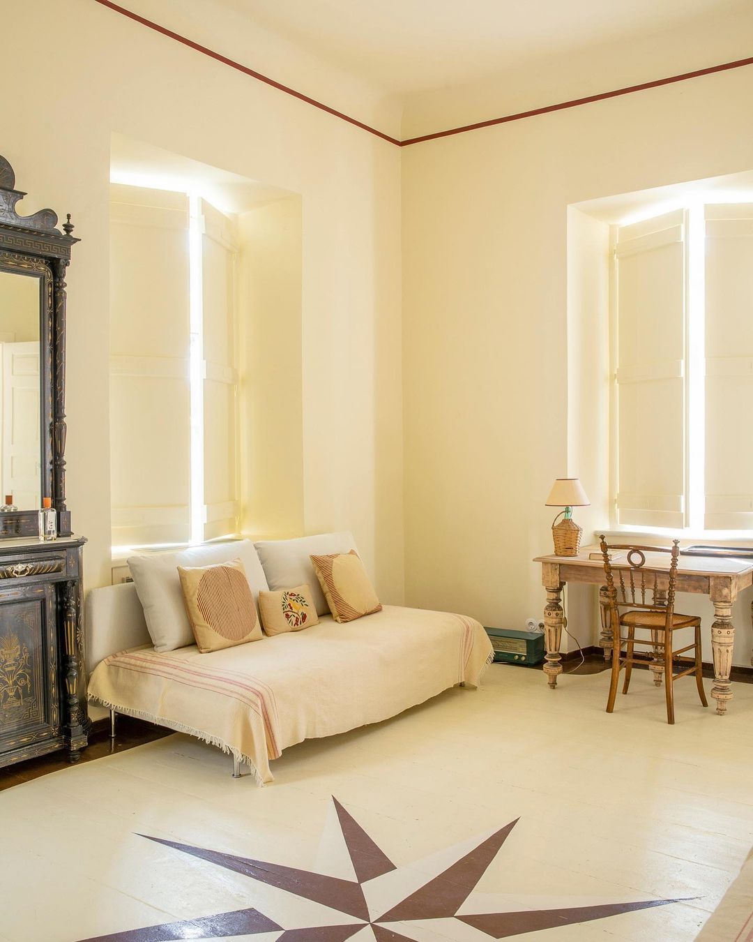 Interior view of a sitting room with daybed, writing desk, chair, antique dresser, large windows with shutters, and star pattern on the floor.
