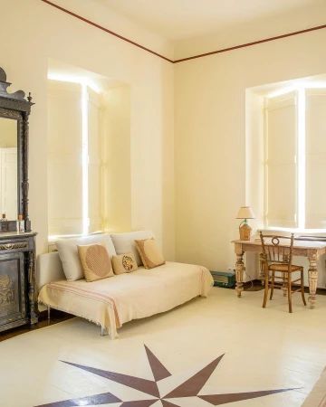 Interior view of a sitting room with daybed, writing desk, chair, antique dresser, large windows with shutters, and star pattern on the floor.
