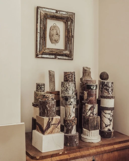 Still life composition featuring stacked marble cylinders and a framed piece of art on a wood dresser against a cream-colored wall.