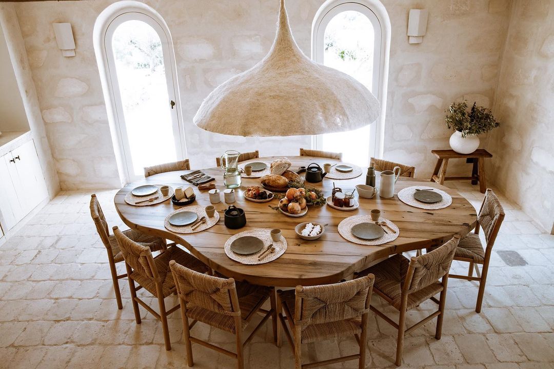 Dining room scene with a long wooden table set for a meal, surrounded by woven chairs, under a large pendant light.