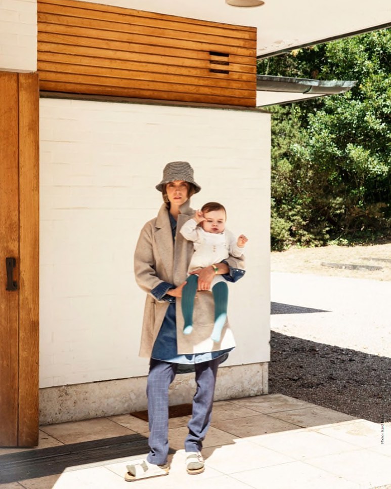 Woman in a tan coat and hat holds a baby with teal tights outside a modern home with wood and white walls and green foliage.