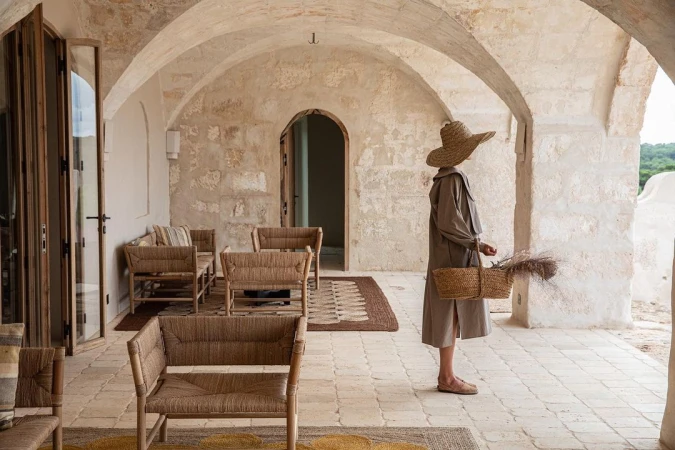 Woman in hat with woven basket on rustic terrace with stone arches and woven furniture, natural light, neutral colors.