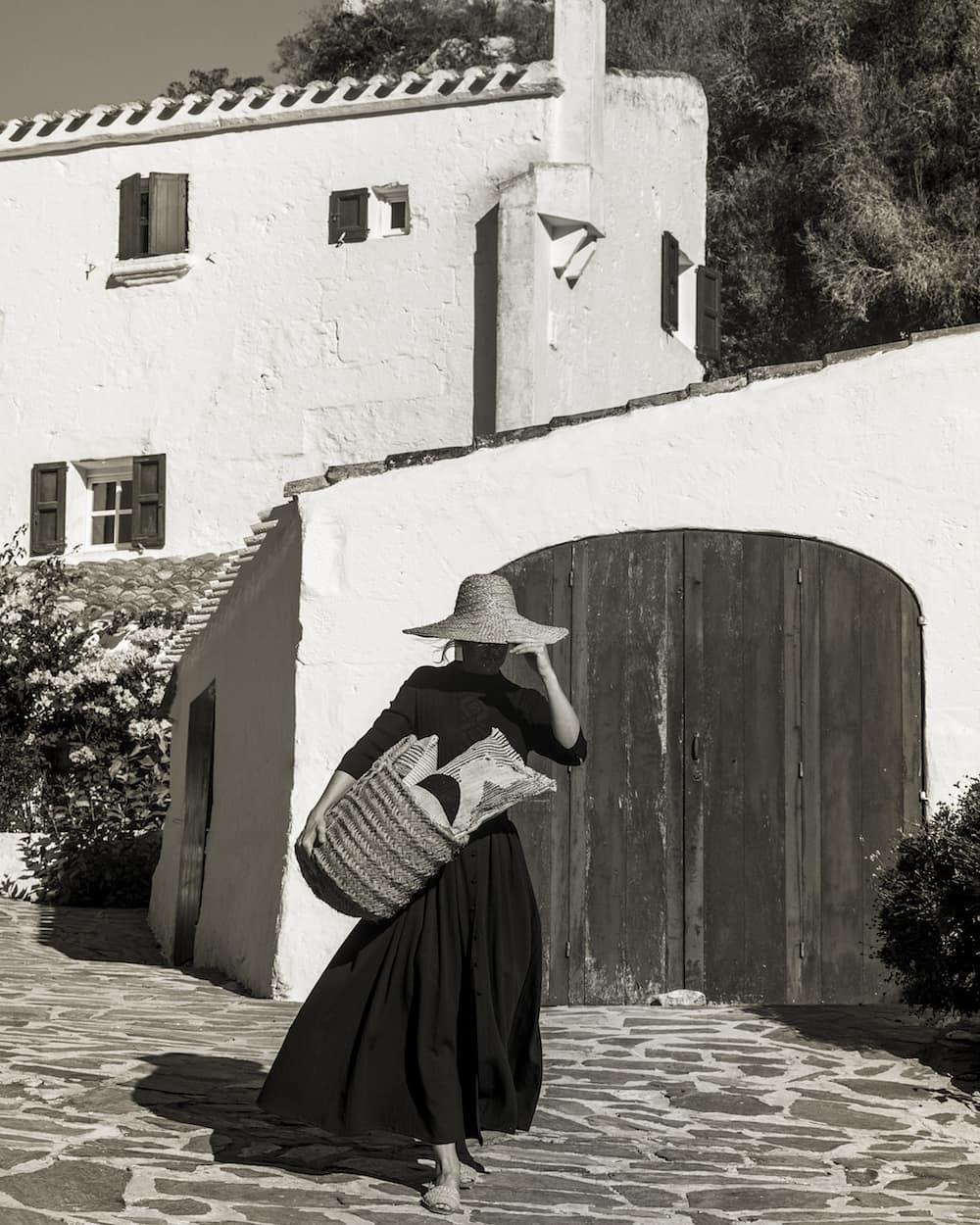 A woman in a black dress and straw hat carries a basket in front of a white building with dark doors and window shutters.