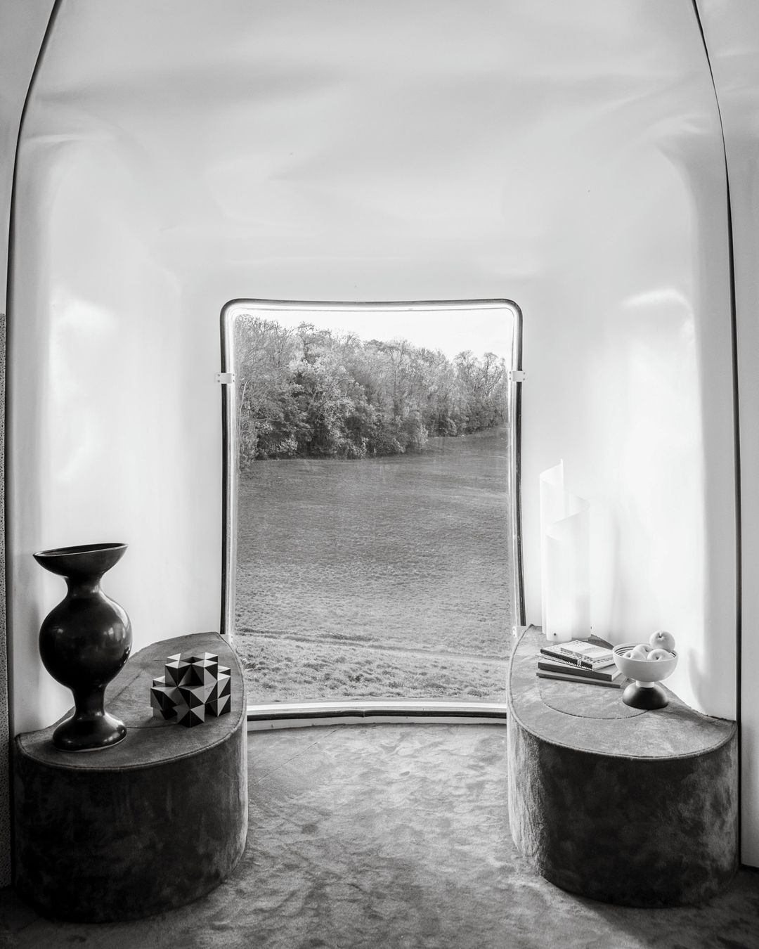 Black and white interior shot with a window overlooking a river and trees, with tables displaying a vase and geometric objects.