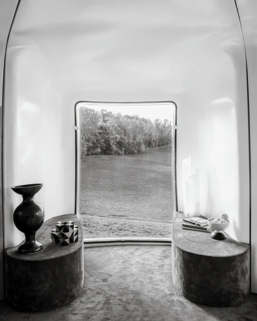 Black and white interior shot with a window overlooking a river and trees, with tables displaying a vase and geometric objects.