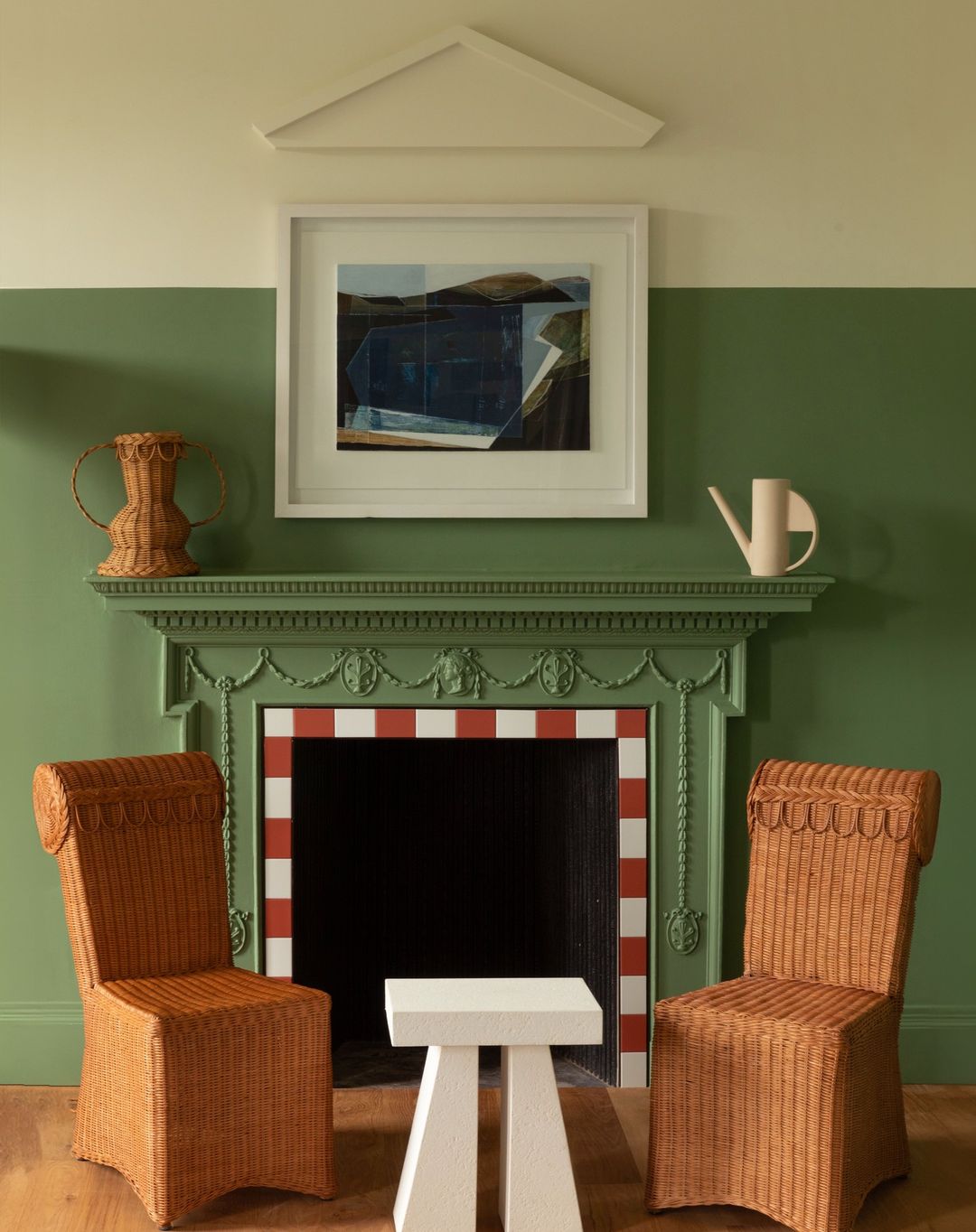 Interior view of a green and white living room with brown wicker chairs flanking a small white table and fireplace mantel with artwork.