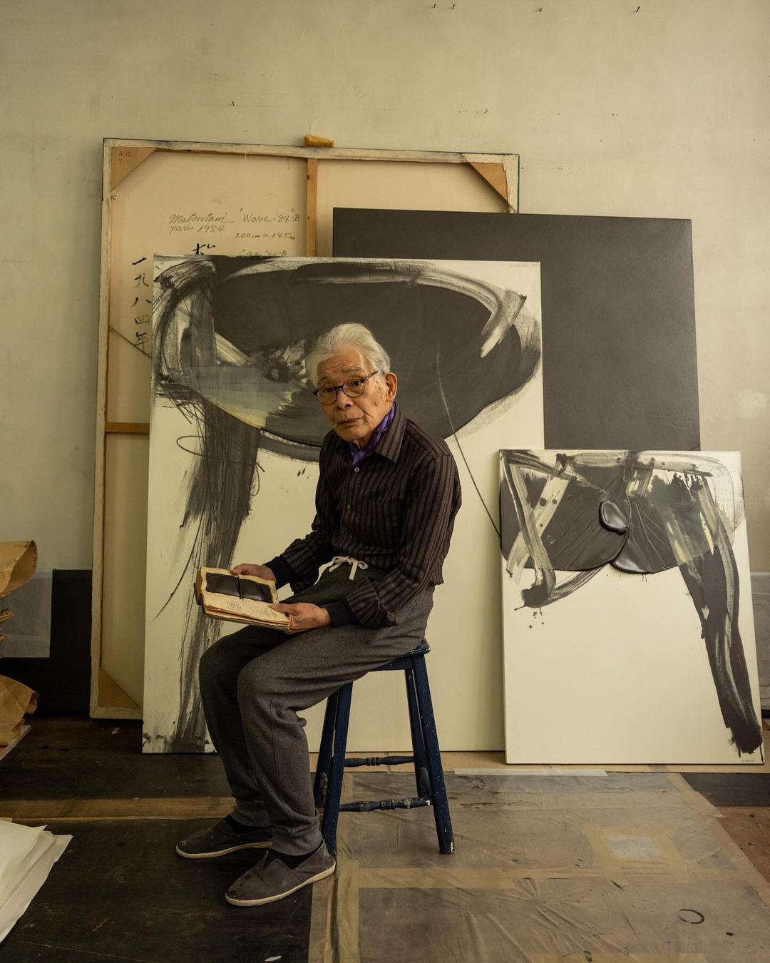 Elderly Asian artist sits in his studio surrounded by his abstract paintings. He is holding an open notebook in his hands.