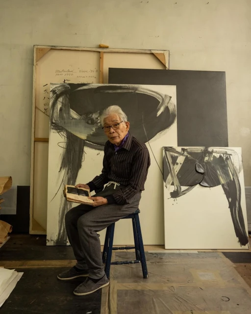 Elderly Asian artist sits in his studio surrounded by his abstract paintings. He is holding an open notebook in his hands.