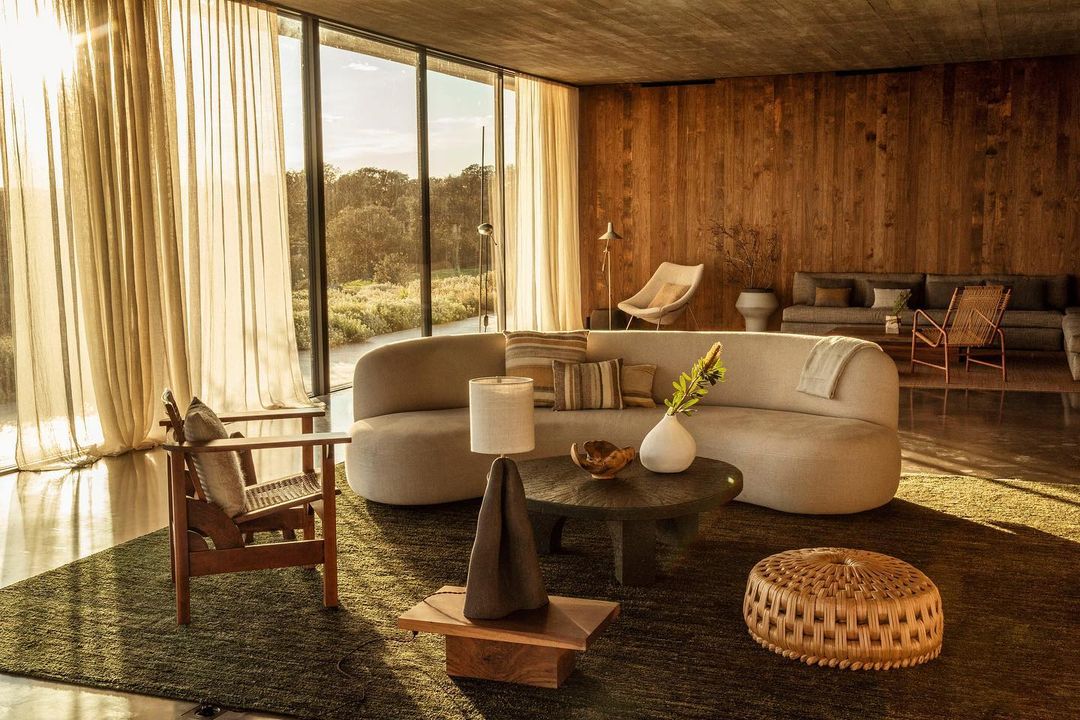 Interior of a living room with modern furniture, including a curved sofa, wooden chairs, a coffee table, and a view to the outside.