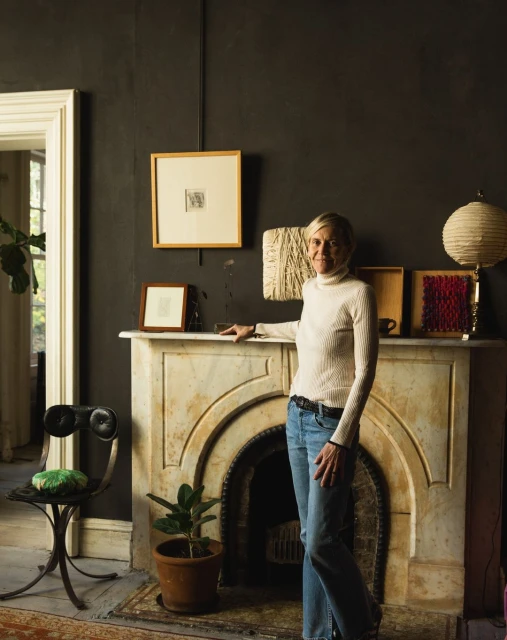 Blond woman in turtleneck sweater stands inside home with gray walls and white marble fireplace, with hanging art and decor.