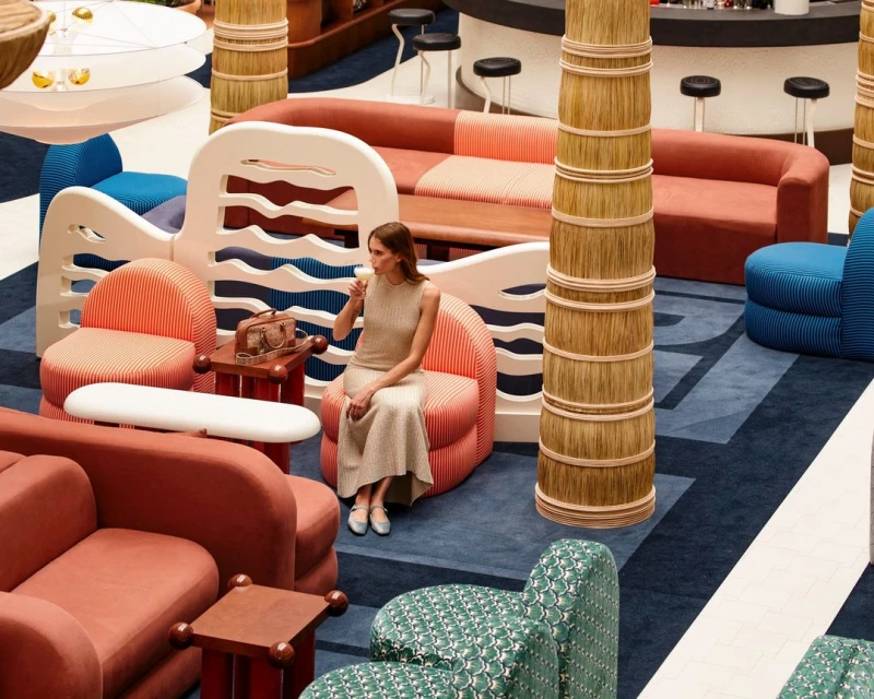 Woman in a neutral dress sits in a coral striped chair with a drink in a stylish lounge area with unique furniture and decor.
