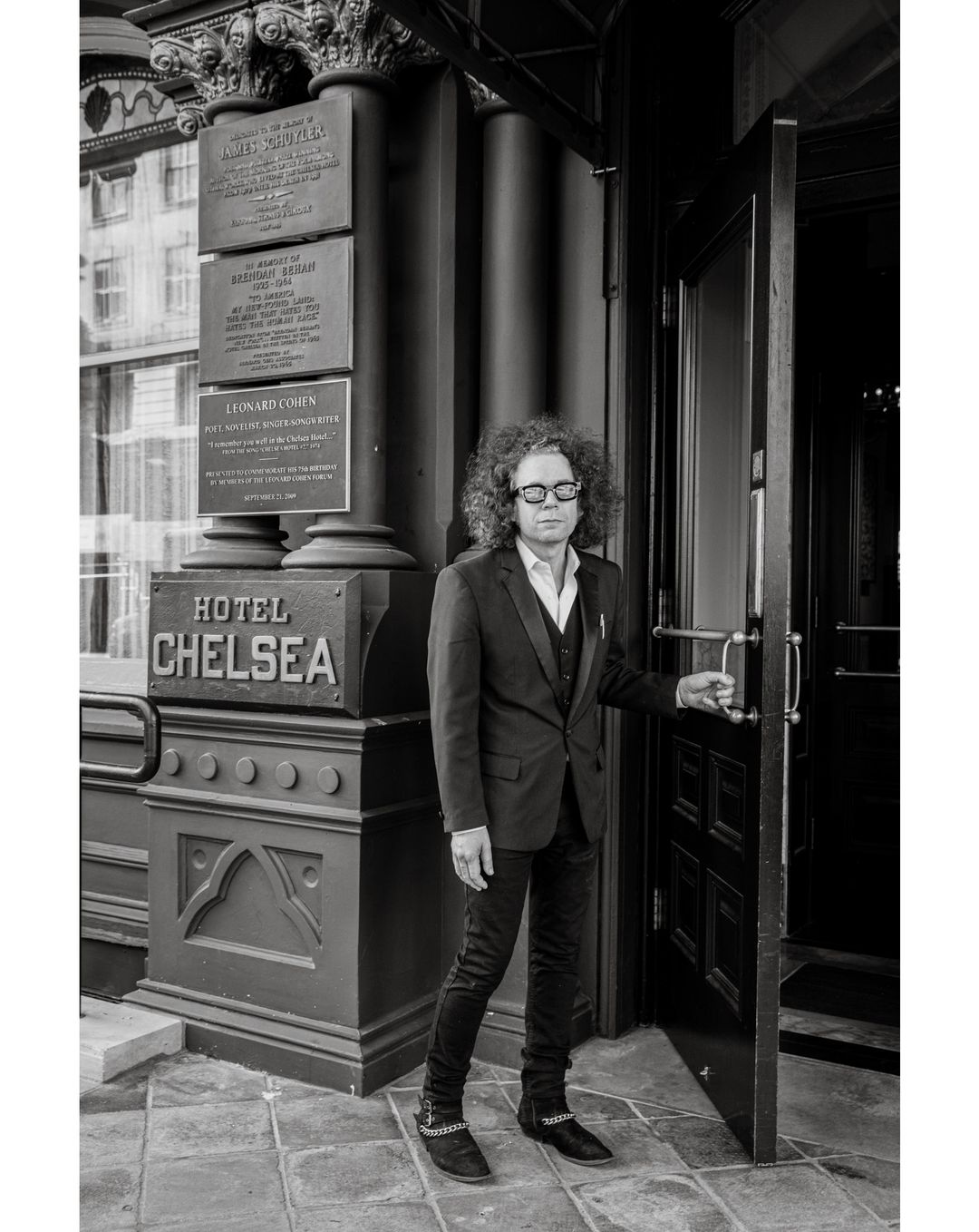 Monochrome portrait of a man in a suit opening the door to the Hotel Chelsea, captured with a medium shot and a neutral expression.