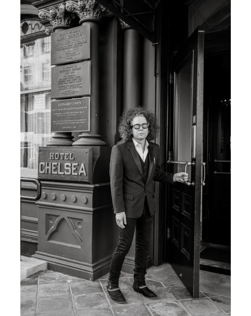 Monochrome portrait of a man in a suit opening the door to the Hotel Chelsea, captured with a medium shot and a neutral expression.