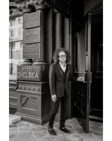 Monochrome portrait of a man in a suit opening the door to the Hotel Chelsea, captured with a medium shot and a neutral expression.