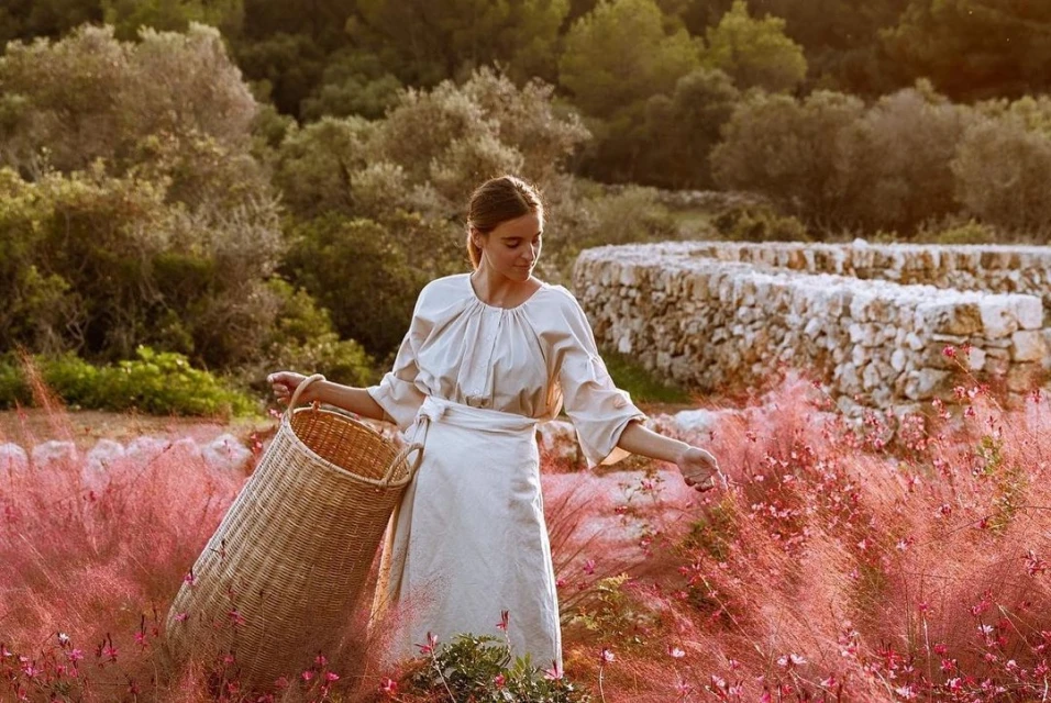 Woman in a light dress walks through a field of pink flowers carrying a wicker basket, with trees and a stone wall in the background.