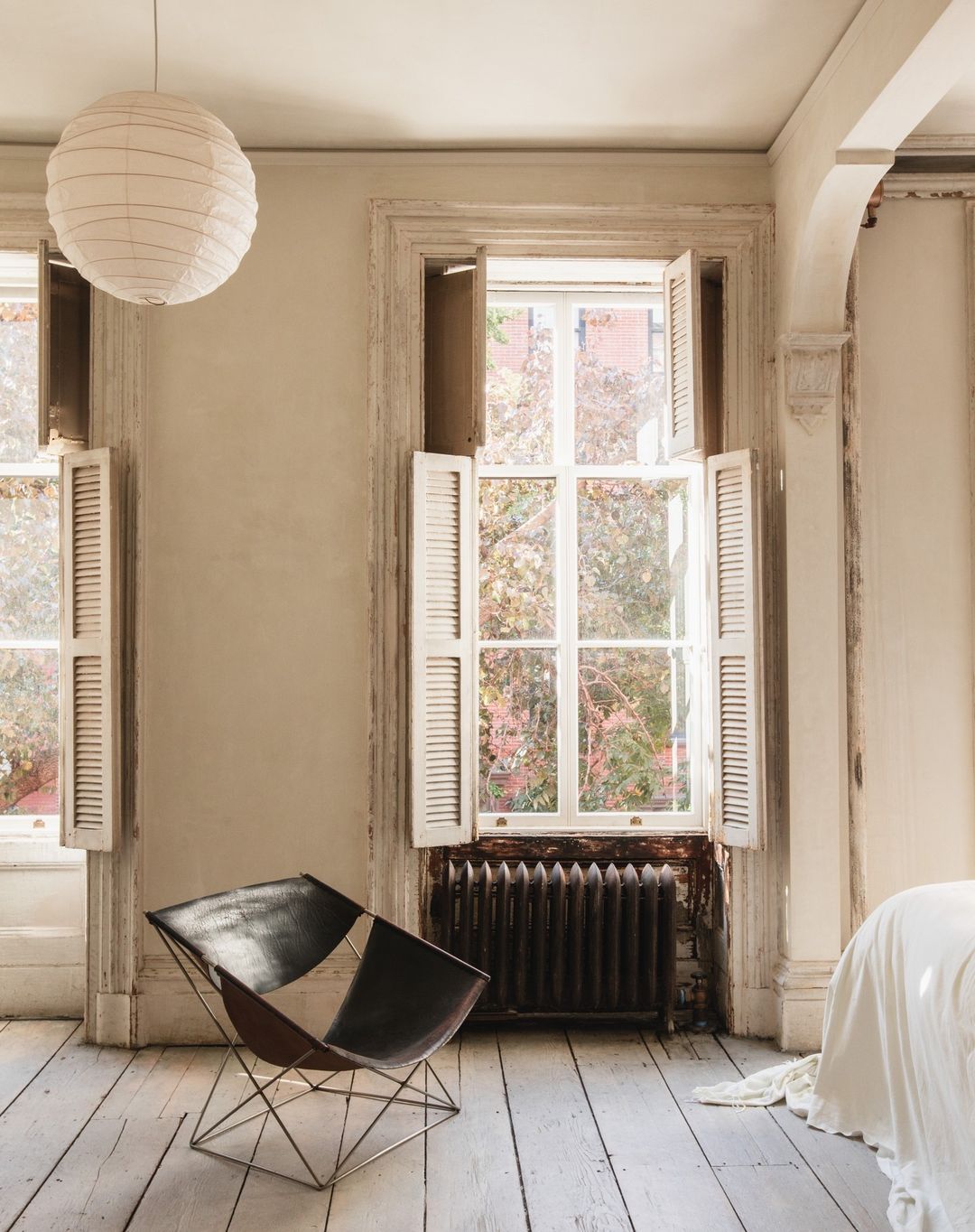 Interior view of a room featuring a modern chair, two windows with white shutters, a radiator, a white paper lantern, and wood floors.