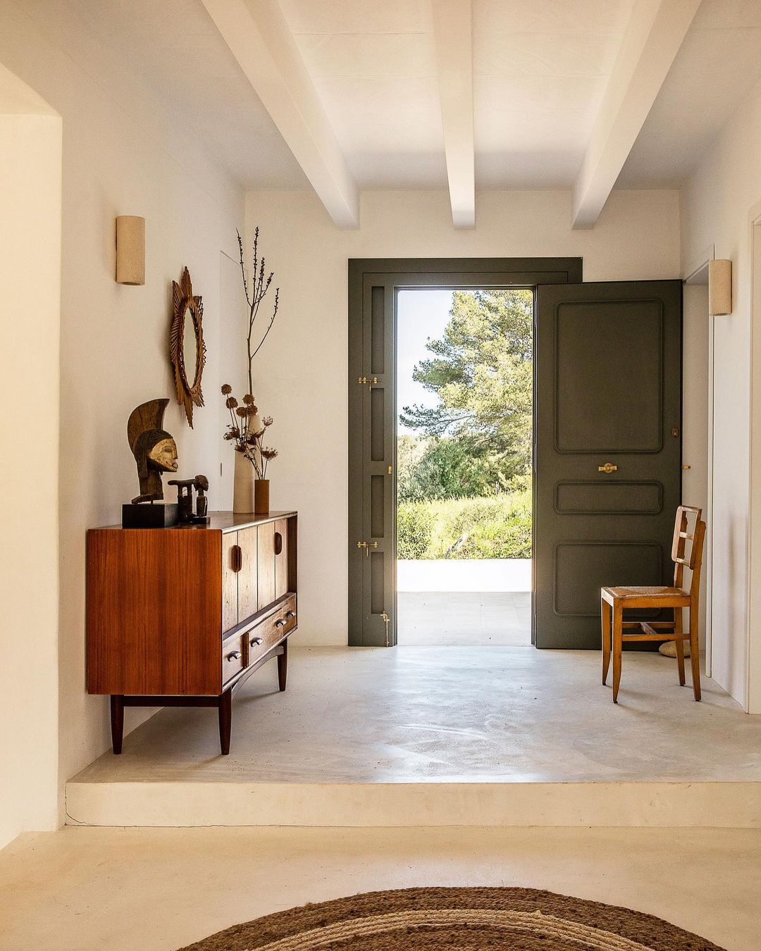 Interior view of a hallway featuring a mid-century modern cabinet, decorative mirror, and an open doorway with a view of trees.