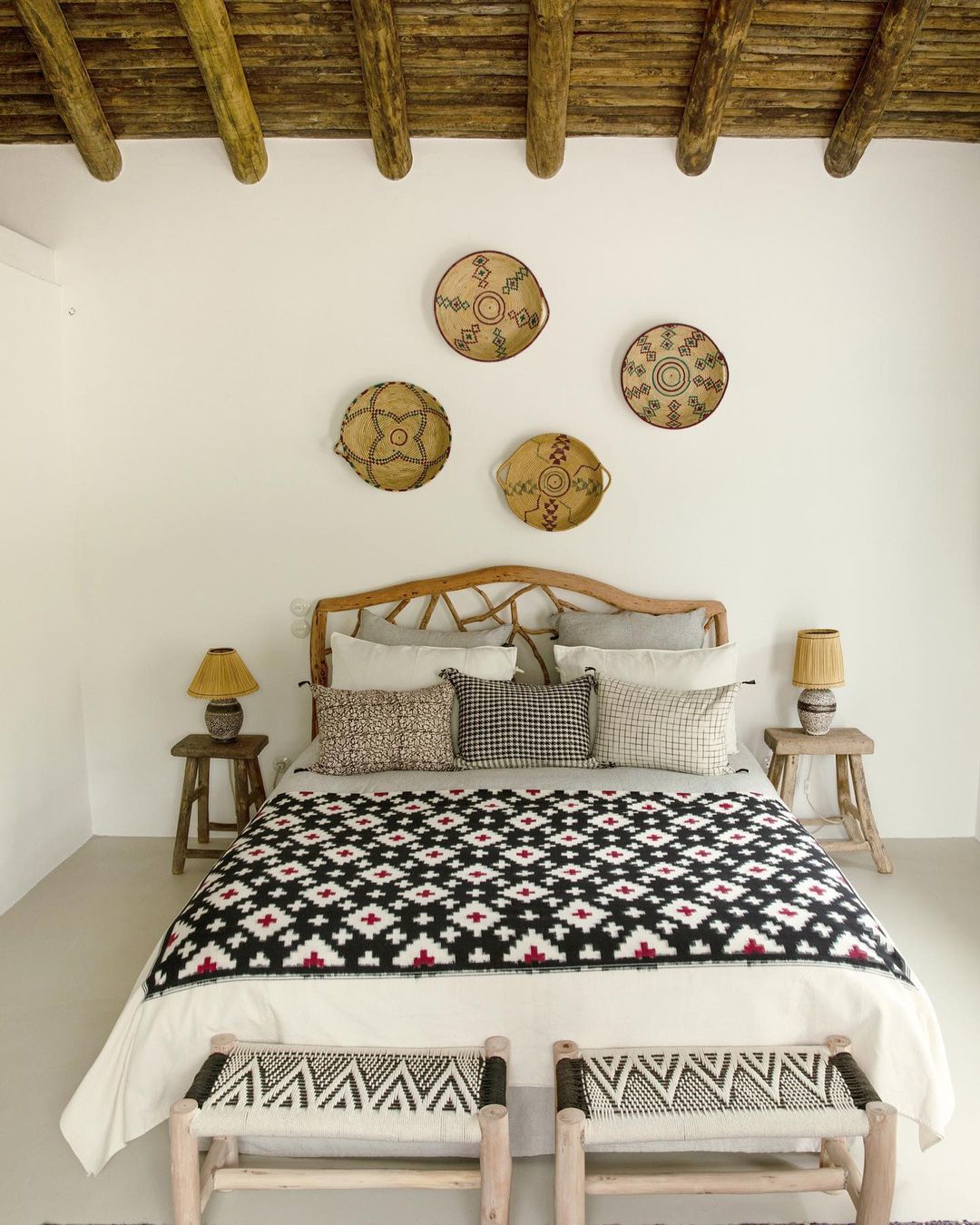 Bedroom interior featuring a bed with a patterned blanket, wooden headboard, and two small wooden stools against a white wall backdrop.