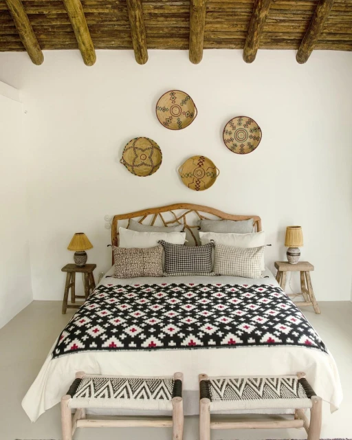 Bedroom interior featuring a bed with a patterned blanket, wooden headboard, and two small wooden stools against a white wall backdrop.