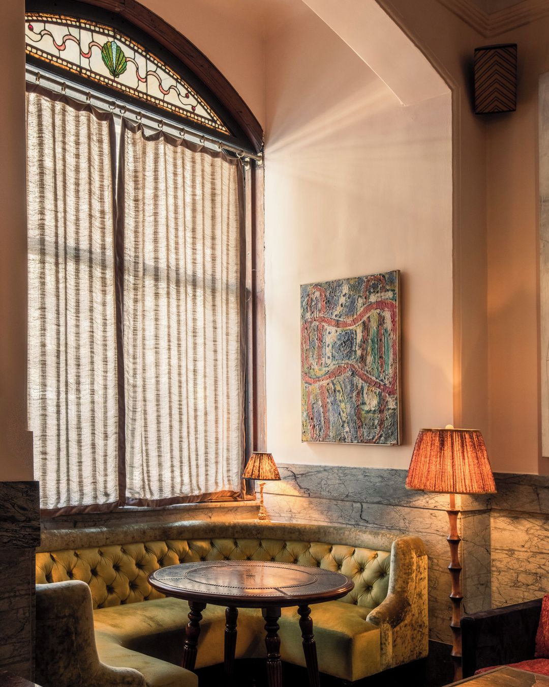 Cozy corner seating area with yellow-green velvet booth, round dark wood table, and patterned lamps in an elegant, vintage-style interior.